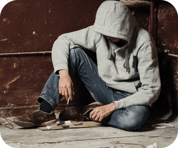 Человек в капюшоне сидит на земле а в руках шприц